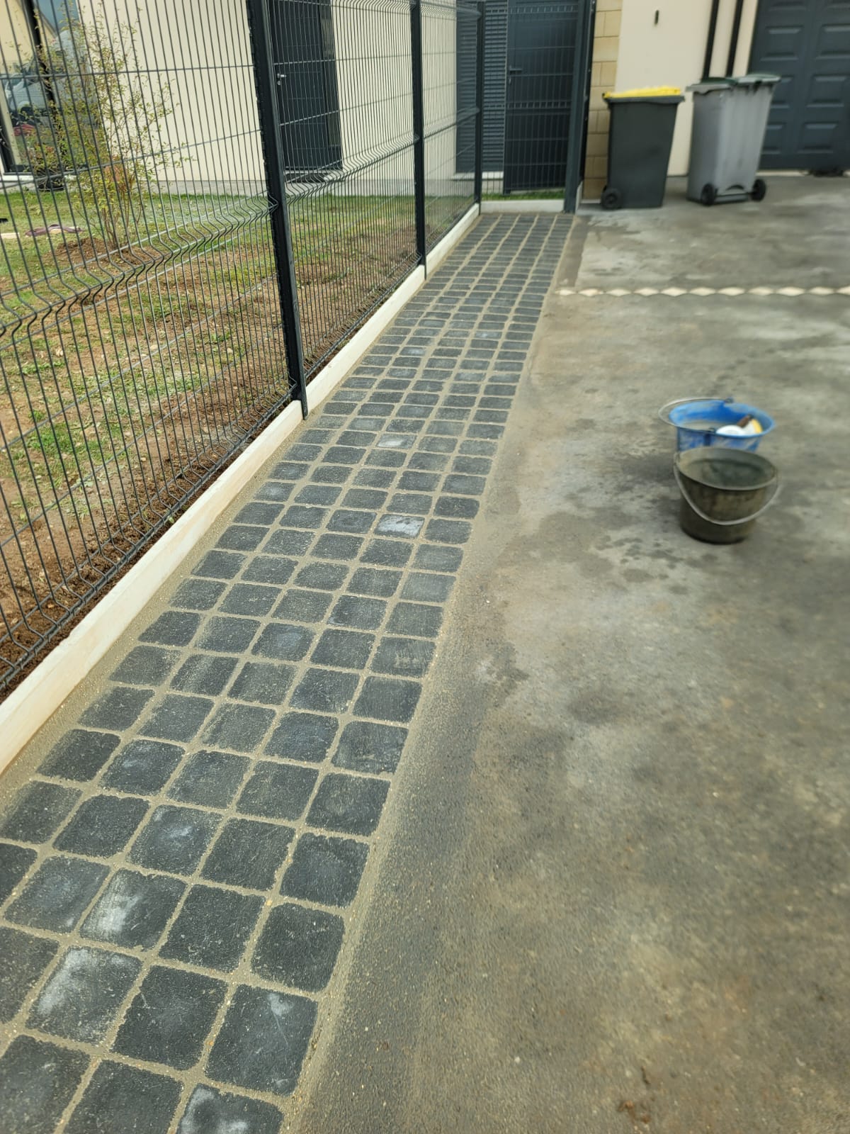 Allee pavee noire avec joints sable le long d une cloture metallique et deux seaux sur beton