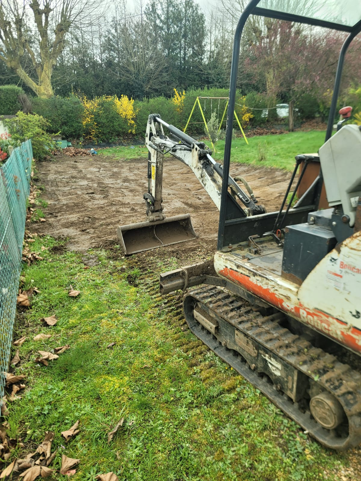 Mini-pelle en action pour terrassement dans un jardin avec pelouse et cloture verte